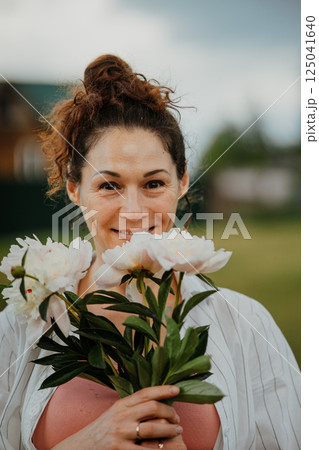 European woman holding bouquet of peonies flowers outdoors near rural house. Female relaxing in backyard garden surrounded by greenery and flowers European woman holding bouquet of peonies flowers outdoors near rural house. Female relaxing in backyard garden surrounded by greenery and flowers 125041640
