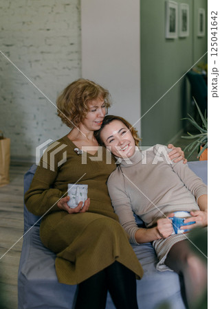 Elderly mother and her daughter relax with tea, expressing their close family bond. The women sit comfortably on a sofa, enjoying a friendly conversation and smiling Elderly mother and her daughter relax with tea, expressing their close family bond. The women sit comfortably on a sofa, enjoying a friendly conversation and smiling 125041642