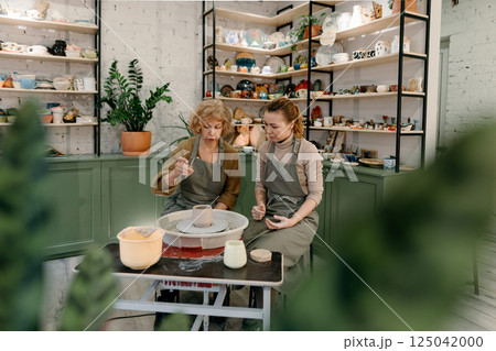 Elderly Caucasian woman practices shaping clay on a pottery wheel at a master class. A young instructor guides her hands, teaching sculpting techniques at a creative workshop 125042000