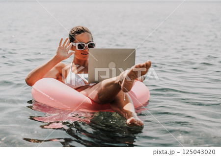 A woman is floating in a pink inflatable raft with a laptop in her hand. She is smiling and she is enjoying her time in the water. 125043020