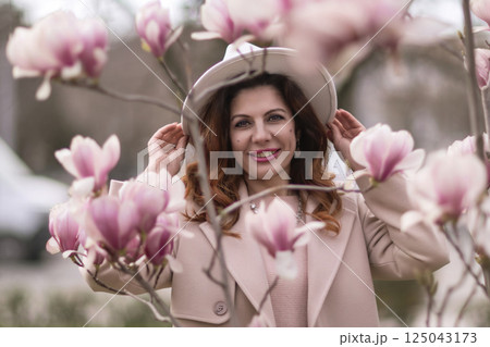 Woman magnolia flowers, surrounded by blossoming trees., hair down, white hat, wearing a light coat. Captured during spring, showcasing natural beauty and seasonal change. 125043173