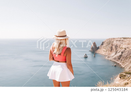 A woman in a pink top and white skirt is standing on a cliff overlooking the ocean. The scene is serene and peaceful, with the woman taking in the beautiful view. A woman in a pink top and white skirt is standing on a cliff overlooking the ocean. The scene is serene and peaceful, with the woman taking in the beautiful view. 125043184
