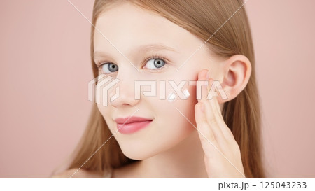 Portrait of a smiling cheerful girl applying face cream on cheek for skin glow. Advertising face cream and skincare products for teens. Blonde girl with blue eyes isolated in studio on pink background 125043233