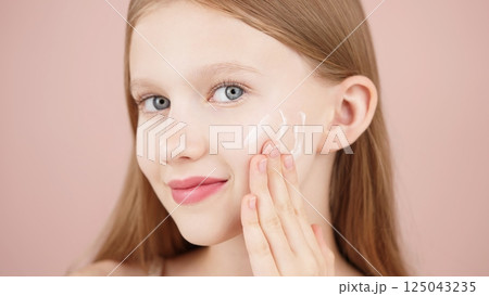 Portrait of a smiling cheerful girl applying face cream on cheek for skin glow. Advertising face cream and skincare products for teens. Blonde girl with blue eyes isolated in studio on pink background 125043235