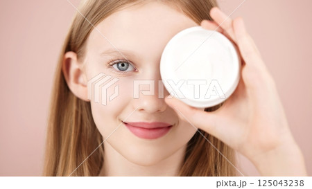 Portrait of a funny smiling blonde young girl posing in studio, holding a cosmetic cream jar in front of her eye on pink background. Advertising face cream. Skincare products for teens or kids 125043238