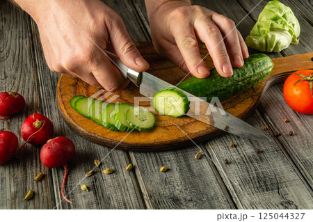 A person skillfully cuts a fresh cucumber into thin slices on a wooden cutting board. Nearby, bright red radishes and a tomato lay, adding color to the rustic kitchen setting A person skillfully cuts a fresh cucumber into thin slices on a wooden cutting board. Nearby, bright red radishes and a tomato lay, adding color to the rustic kitchen setting 125044327