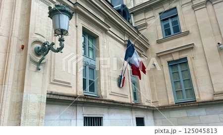 Waving national flags of France and the shield with letters RF Republique Francaise or French Republic on the typical administrative building Waving national flags of France and the shield with letters RF Republique Francaise or French Republic on the typical administrative building 125045813