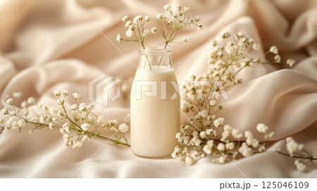 Close-up view of a glass bottle of milk surrounded by delicate flowers on a light beige fabric backdrop in a studio setting 125046109