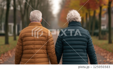 Elderly couple walking hand in hand down a serene path, surrounded by autumn foliage, showcasing companionship and love in a peaceful outdoor setting. 125050383