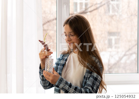 Girl holding small glass vase with flowers near window. 125054932