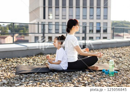 Caucasian adult woman and young girl sitting back-to-back meditating outdoors together on a rooftop. They practice yoga and mindfulness during a sunny day. Caucasian adult woman and young girl sitting back-to-back meditating outdoors together on a rooftop. They practice yoga and mindfulness during a sunny day. 125056845
