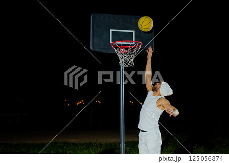 A man performs a slam dunk on a basketball court at night under the glare of bright outdoor lights 125056874