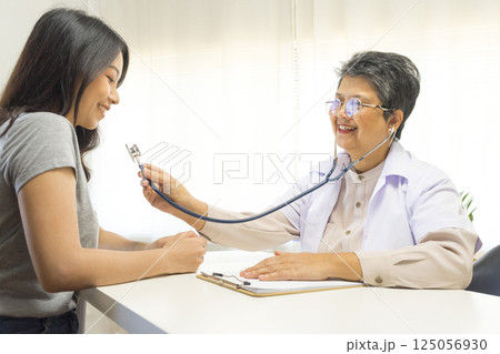 Smiling mature female general practitioner sitting at table and talking to patient Smiling mature female general practitioner sitting at table and talking to patient 125056930
