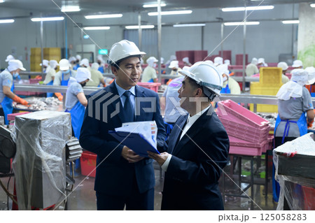 Two factory managers in safety helmets discussing documents inside a busy canned fish processing plant Two factory managers in safety helmets discussing documents inside a busy canned fish processing plant 125058283