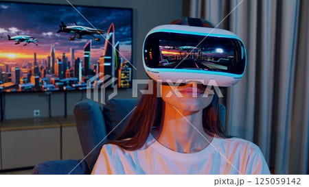 Immersed in virtual reality, a woman explores a futuristic cityscape with flying vehicles, enjoying a simulated experience from her comfortable chair at home 125059142