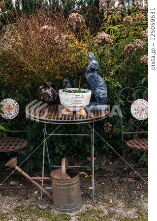 Vintage garden table with mosaic chairs, ceramic planter labeled LE JARDINIER, and whimsical animal sculptures surrounded by autumn foliage. Rustic garden decor, slow living Vintage garden table with mosaic chairs, ceramic planter labeled LE JARDINIER, and whimsical animal sculptures surrounded by autumn foliage. Rustic garden decor, slow living 125059631