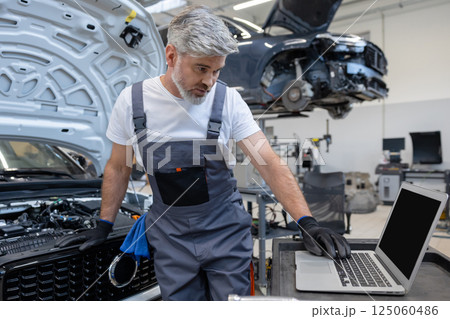 Man technician looking at inspection vehicle details under car hood using laptop for diagnostic Man technician looking at inspection vehicle details under car hood using laptop for diagnostic 125060486