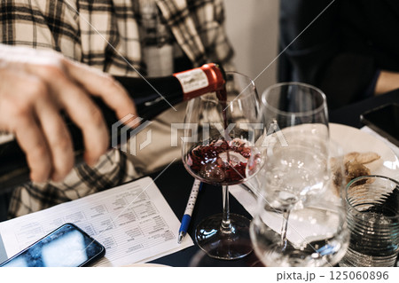 Participant pouring red wine into a glass during a wine workshop session with a tasting card nearby. Sommelier training, hands-on wine education, sensory learning, grape varietals 125060896