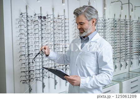 Mature doctor in lab coat standing near the stand with glasses and looking confident Mature doctor in lab coat standing near the stand with glasses and looking confident 125061209