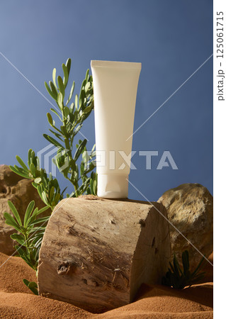 Unbranded white cream tube stands on a raw wooden log, the wood surface shows natural scratches, with rough, fibrous textures along the edge. Rough and uneven rocks displayed on the sand field. Unbranded white cream tube stands on a raw wooden log, the wood surface shows natural scratches, with rough, fibrous textures along the edge. Rough and uneven rocks displayed on the sand field. 125061715