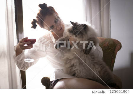 Woman holding a fluffy cat and drinking red wine by the window 125061904