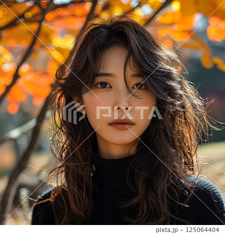 Woman with long brown hair is standing in front of a tree with orange leaves 125064404