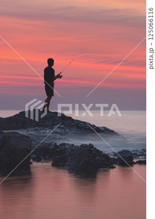 Sea fishing in the tropics. The silhouette of a fisherman on the rocks among the blurred waves and water with red highlights from the sunset. Stunning seascape 125066116