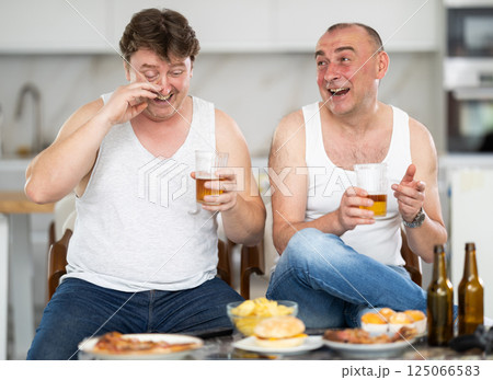 Two adult male friends drinking beer and eating chips at home. Bad habits, eating disorder, alcoholism Two adult male friends drinking beer and eating chips at home. Bad habits, eating disorder, alcoholism 125066583