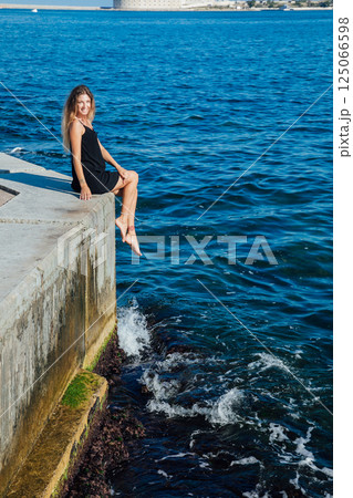 Woman in black dress sitting on pier and looking at the sea 125066598