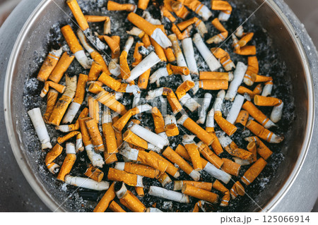 Close-up top view discarded cigarette butts filling ashtray, showcasing smoking addiction and toxic waste. Heavy concentration of nicotine residue and ashes threatens environment and personal health 125066914