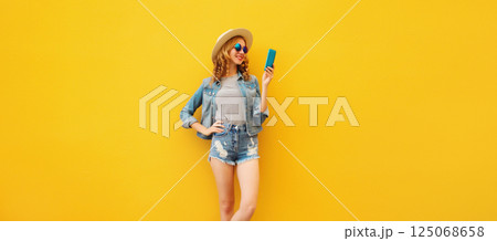 Summer stylish happy young woman with smartphone in straw tourist hat posing on yellow background Summer stylish happy young woman with smartphone in straw tourist hat posing on yellow background 125068658
