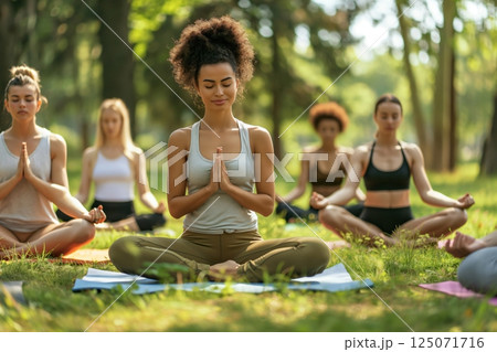 Group of women are sitting in a park on yoga mats with their legs crossed, hands pressed together and eyes closed, meditating 125071716