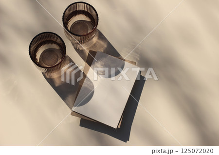 Summer neutral wedding stationery. Closeup of blank greeting card, envelope branding mockup. Two glass of water of in sunlight, long shadows. Invitation, template. Beige table background. Flatlay, top 125072020