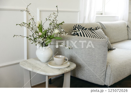 Springtime breakfast still life. Cup of coffee tea, marble table. Tulips blooming cherry plum tree branches bouquet in vase. Blurred cozy linen sofa with cushions. Elegant living room view. Home decor 125072026