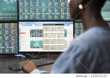 Closeup of black female employee typing on a keyboard at office desk, reviewing stock exchange progress on multiple screens. Selective focus on digital monitor displaying trading statistics and data. 125072127