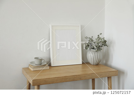 Beautiful Scandinavian interior. Green dry eucalyptus tree branches in vase on desk, table. Cup of coffee tea, old books. Empty wooden vertical picture frame mockup. Wall background. Elegant indoor. Beautiful Scandinavian interior. Green dry eucalyptus tree branches in vase on desk, table. Cup of coffee tea, old books. Empty wooden vertical picture frame mockup. Wall background. Elegant indoor. 125072157