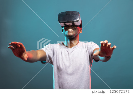 Happy male model enjoying an interactive game with a VR headset, reaching out toward the camera. Excited black man experiencing cutting edge 3D technology for virtual entertainment. Happy male model enjoying an interactive game with a VR headset, reaching out toward the camera. Excited black man experiencing cutting edge 3D technology for virtual entertainment. 125072172