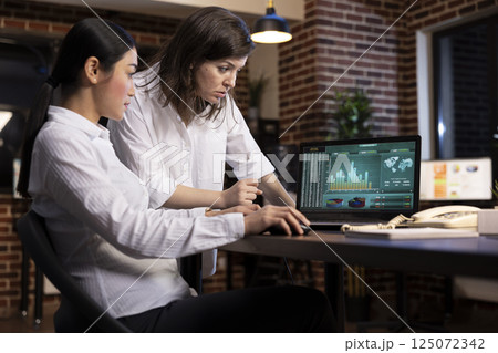 A professional discussion between coworkers, collaborating on a business project, using research data on laptop. White woman stands and reviews details as her colleagues scrolls through the document. 125072342