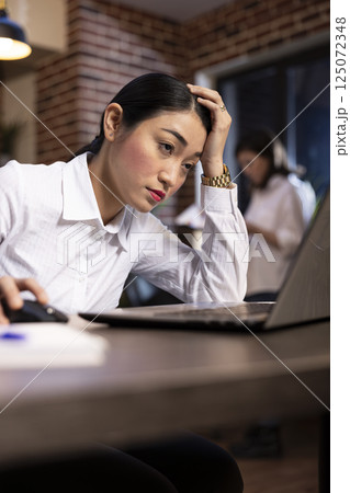 Asian female manager showing signs of fatigue while working late at her desk. Stressed businesswoman leans on one hand and looks at her laptop, as her blurred coworker reviews documents behind her. Asian female manager showing signs of fatigue while working late at her desk. Stressed businesswoman leans on one hand and looks at her laptop, as her blurred coworker reviews documents behind her. 125072348