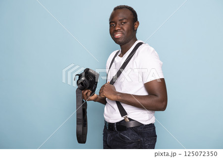 Black man dressed in a casual outfit with suspenders poses with his camera in front of a blue backdrop. Portrait of a male photographer, ideal for advertising, branding, and promotional use. Black man dressed in a casual outfit with suspenders poses with his camera in front of a blue backdrop. Portrait of a male photographer, ideal for advertising, branding, and promotional use. 125072350