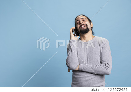 Cheerful young adult laughing and enjoying discussion on a phone call, talking to his friends on a smartphone line against blue background. Smiling person with long hair and lively personality. 125072421