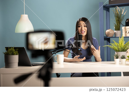 Young female vlogger seated at desk, talking into her podcast mic about marketing strategy for her channel. Asian woman using a camera on tripod, creating lifestyle content for her online audience. Young female vlogger seated at desk, talking into her podcast mic about marketing strategy for her channel. Asian woman using a camera on tripod, creating lifestyle content for her online audience. 125072423