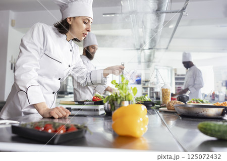 Dedicated chef testing the flavors of a gourmet dish at the kitchen counter, adjusting the seasoning for the perfect taste. Woman focusing on fresh ingredients for fine dining presentation. 125072432