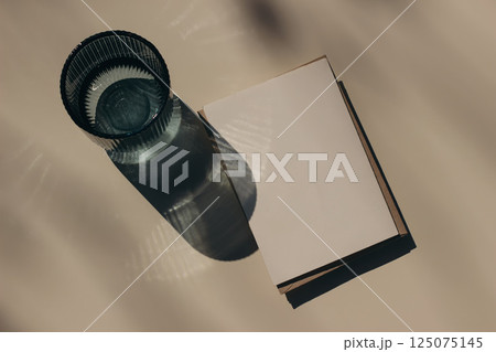 Summer neutral wedding stationery. Closeup of blank greeting card, envelope branding mockup. Blue glass of water in sunlight, long shadows. Invitation, template. Beige table background. Flatlay, top 125075145