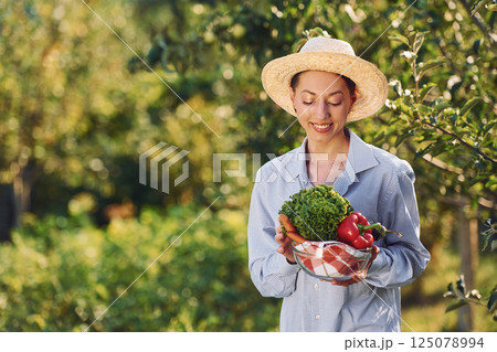 With vegetables in hands. Young cheerful woman is in the garden at daytime 125078994