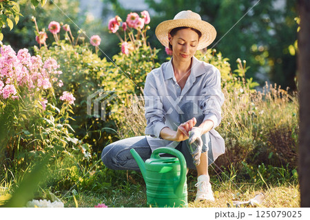 Preparing for watering the flowers. Young cheerful woman is in the garden at daytime Preparing for watering the flowers. Young cheerful woman is in the garden at daytime 125079025