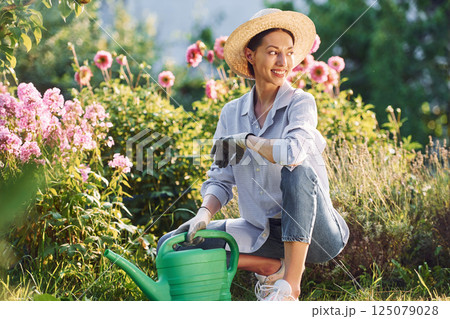 Preparing for watering the flowers. Young cheerful woman is in the garden at daytime Preparing for watering the flowers. Young cheerful woman is in the garden at daytime 125079028