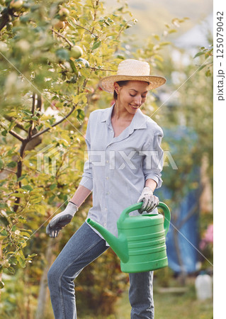 Standing and holding watering can. Young cheerful woman is in the garden at daytime Standing and holding watering can. Young cheerful woman is in the garden at daytime 125079042