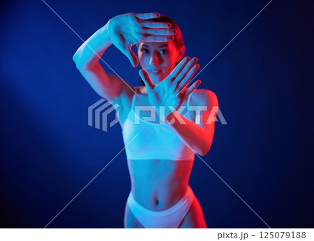 Doing gestures by the hands. Young woman in underwear is in the studio with neon lights 125079188