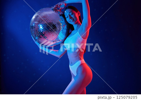 With shiny mirrored ball. Young woman in underwear is in the studio with neon lights With shiny mirrored ball. Young woman in underwear is in the studio with neon lights 125079205